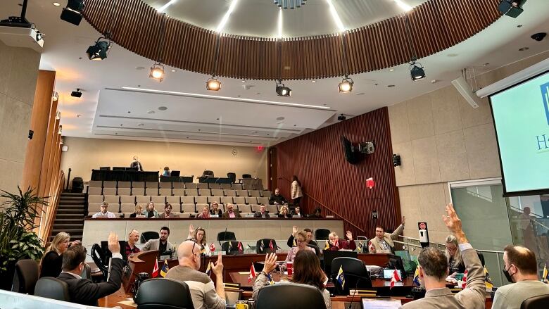 People sitting around a horseshoe-shaped table in a city council chamber raise their hands. 