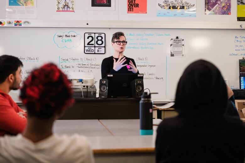 A teacher stands speaking at the front of a classroom, with a whiteboard filled with notes behind them.