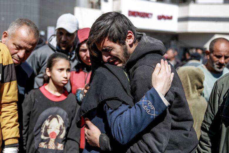 A bearded man with an anguished look on his face embraces a person whose face is not visible. Both are standing in front of a crowd of people outdoors.