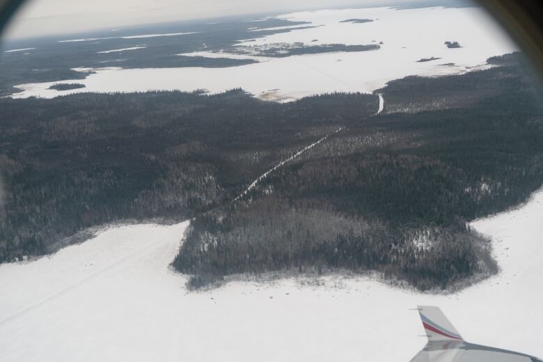 An aerial shot, taken through a plane window, of a winter road stretching through a wooded area across two snow-covered bodies of water.