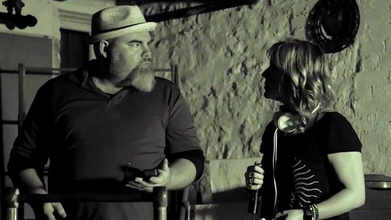 Black and white image of bearded man (left) in hat looking at woman at right. Woman is wearing short-sleeved shirt and has headphones around her neck. They are standing in front of a stone wall inside a basement. 