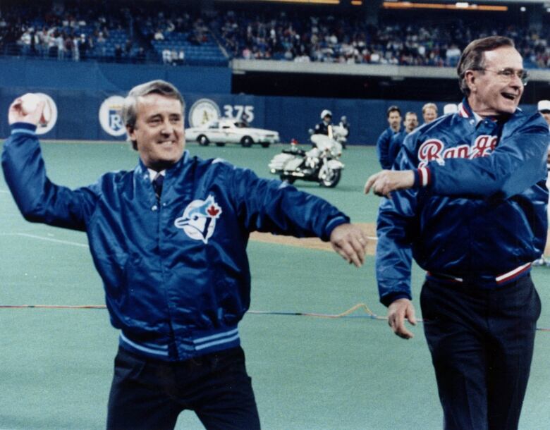 Two men in baseball jackets throw baseballs on a large porfessional field.