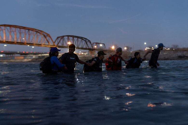 Silhouette of people wading, stomach-deep, through a river at night, with bridge in the background