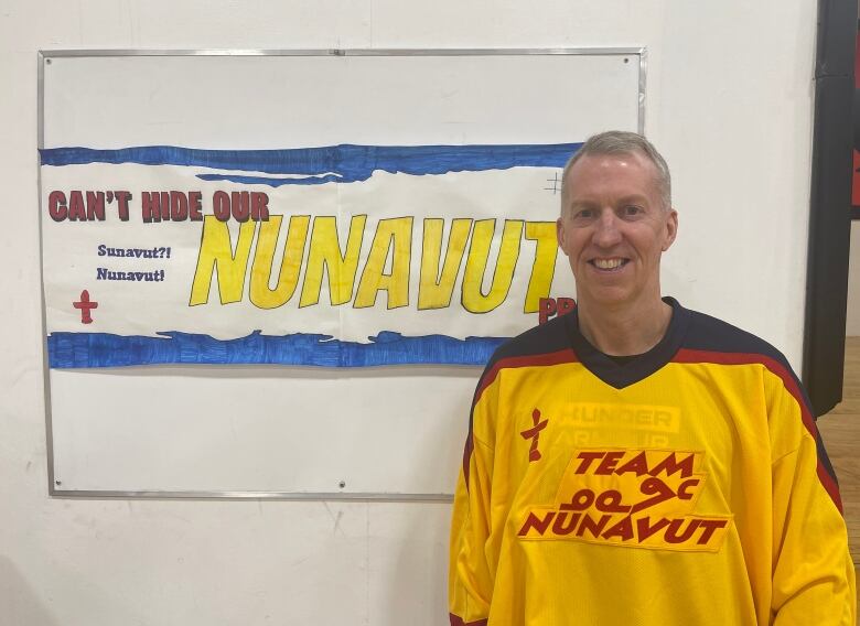 A man in a yellow 'Team Nunavut' jersey stands in front of a poster on a wall.