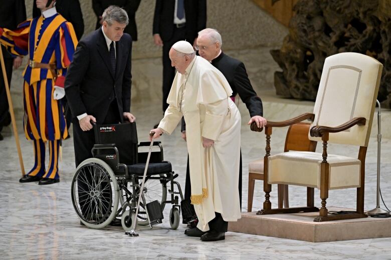 A man in white and a white skullcap leans on a cane. Two other man stand nearby, one with an empty wheelchair.