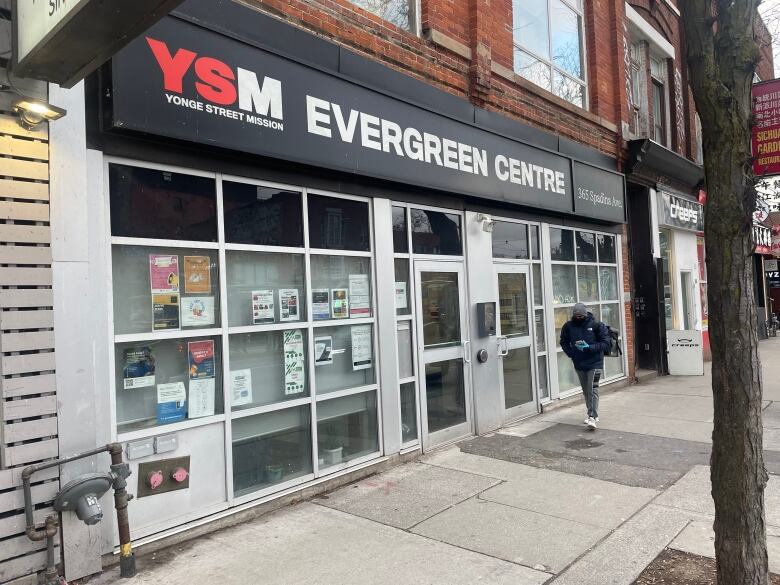 The facade of a cement building reading Y-M-S Evergreen Centre. The day is grey. The building is on a city sidewalk. A pedestrian passes the building