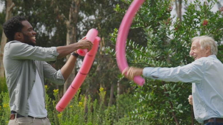 Two men flight playfully with pink pool noodles. One noodle is thicker than the other.