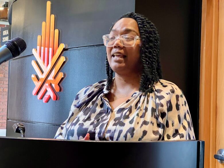 A woman wearing glasses and a black and white print blouse stands behind a podium.