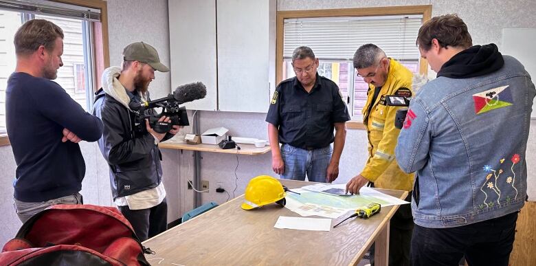 Members of a film crew are standing at a table filming two firefighters who are referencing to a yellow helmet and paperwork on the table. 