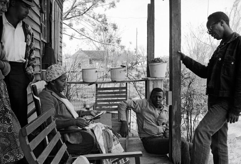 In a black and white photo, a group of Black men and women gather on a porch and are having a conversation. 