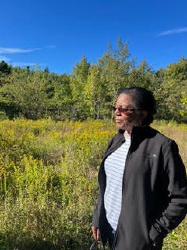 A Black woman wearing sunglasses stands in a field under a blue sky.