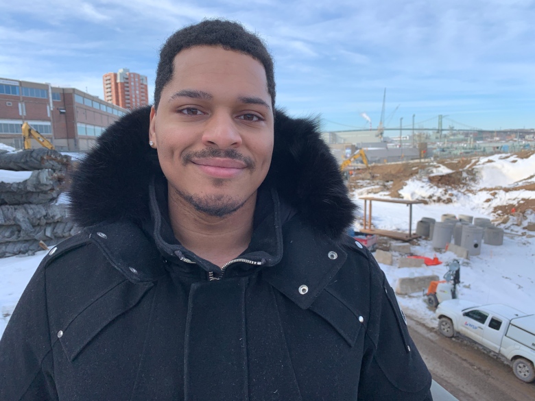 A young Black man in a black winter jacket stands beside a construction zone.