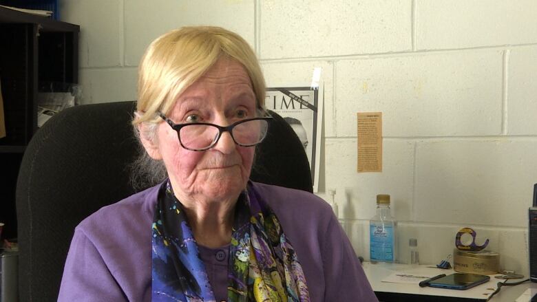 A woman with glasses and wearing a purple jacket sits in an office while doing an interview. 