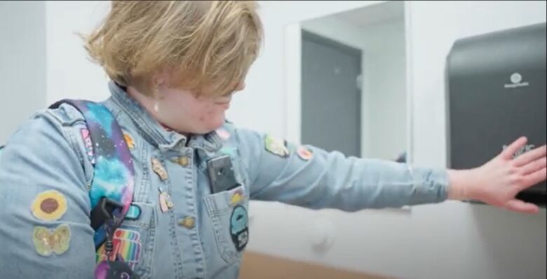 A woman with short blond hair and wearing a denim jacket holds her hand under a hand dryer in a washroom.