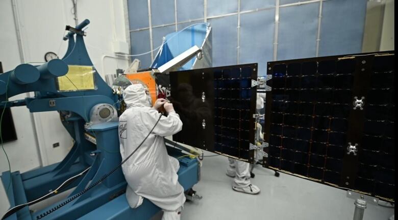 Workers dressed head-to-toe in protective gear make adjustments to the solar panels on a methane-hunting satellite. 