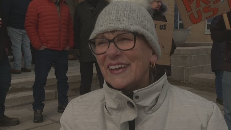 A woman wearing a winter hat and glasses smiles in front of protestors. 