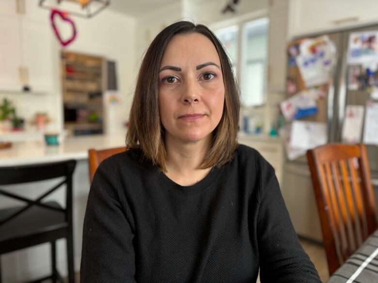 A woman sits at a table with a kitchen, out of focus, behind her.