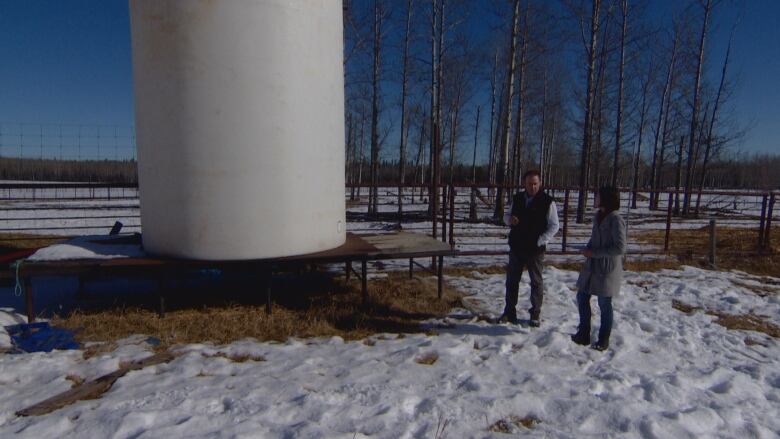 A man and woman stand beside a large water tank on a snow-covered ranch.