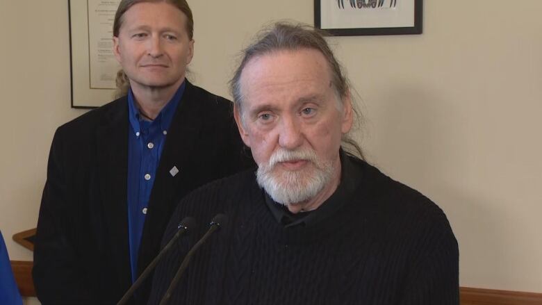 A man with a grey beard is wearing a black shirt and speaking at a podium. Behind him is a man in a blue shirt and black suit. 