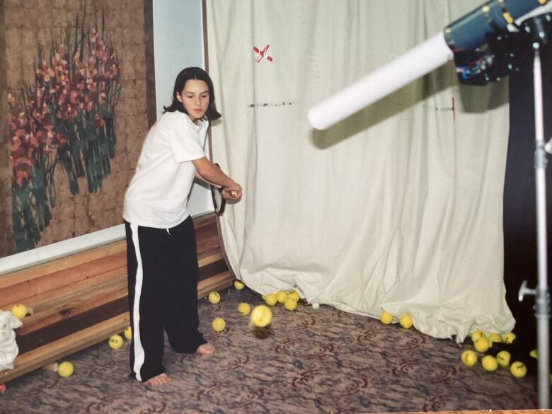 A young Gaby Dabrowski holds a racket, ready to hit strokes at the yellow tennis balls approaching her from a tube like machine to the right of the frame.