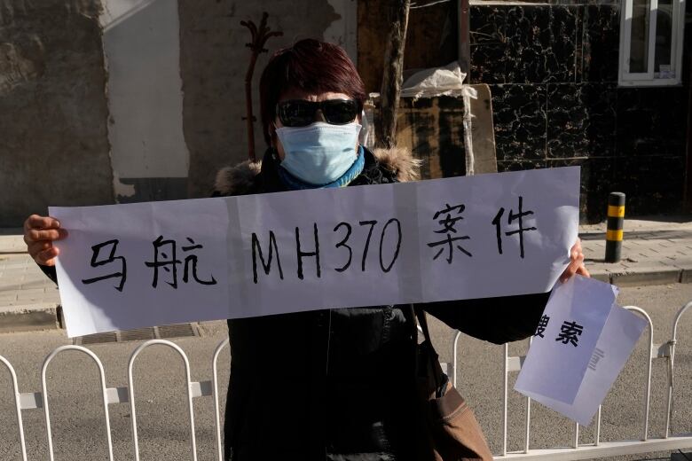 A woman in a mask and sun glasses holds a sign. 