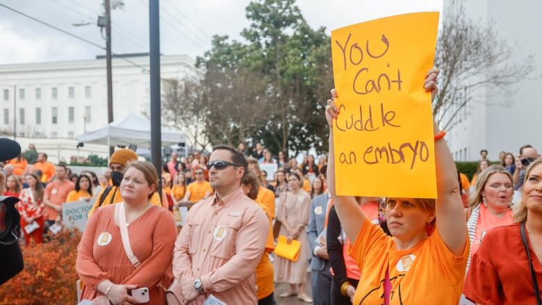 Several people stand outside, all wearing the same colour, in a demonstration. One woman holds up a sign that says, 'You can't cuddle an embryo.'