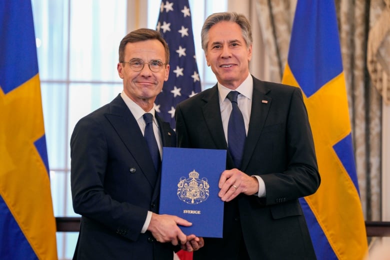 Two cleanshaven men wearing suit and tie, one of them bespectacled, pose for a photo while holding a leather-bound book together.