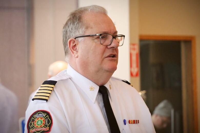 A man with grey hair and glasses wears a black tie and a white uniform shirt with black and gold epaulettes and a fire department badge on the shoulder.