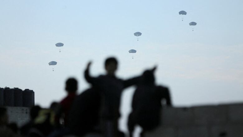 Packages attached to parachutes fall onto a city. Silhouettes of people standing and watching are visible in the foreground.