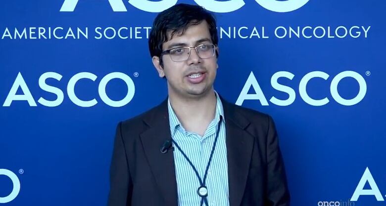 A man stands wearing glasses, a striped shirt, suit jacket and conference lanyard in front of a blue backdrop reading American Society of Clinical Oncology. 