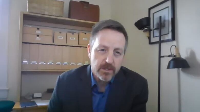 Man wearing a suit sits near a shelf.