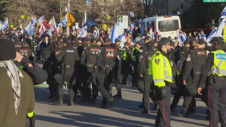 Police officers are seen on a street with protesters, who are shown with Israel and Canada flags.