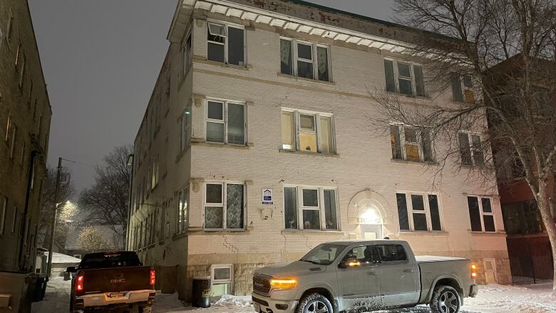 The exterior of a white brick apartment building, with some windows boarded up, is shown.