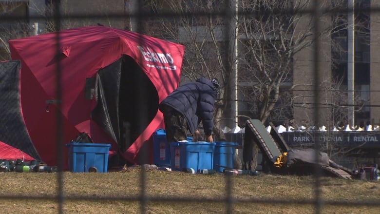 A person behind a fence places a lid on a blue box next to a red tent.