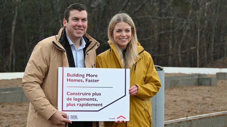 A white woman with long blonde hair in a yellow rain jacket stands outside beside a white man with brown hair and tan jacket. They both hold a small white sign with the slogan 'building more homes, faster'