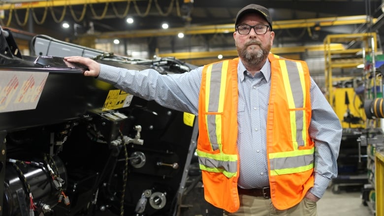 A man in a blue shirt, khakis, a baseball cap and glasses poses next to a header, a piece of farm equipment used to harvest grain.