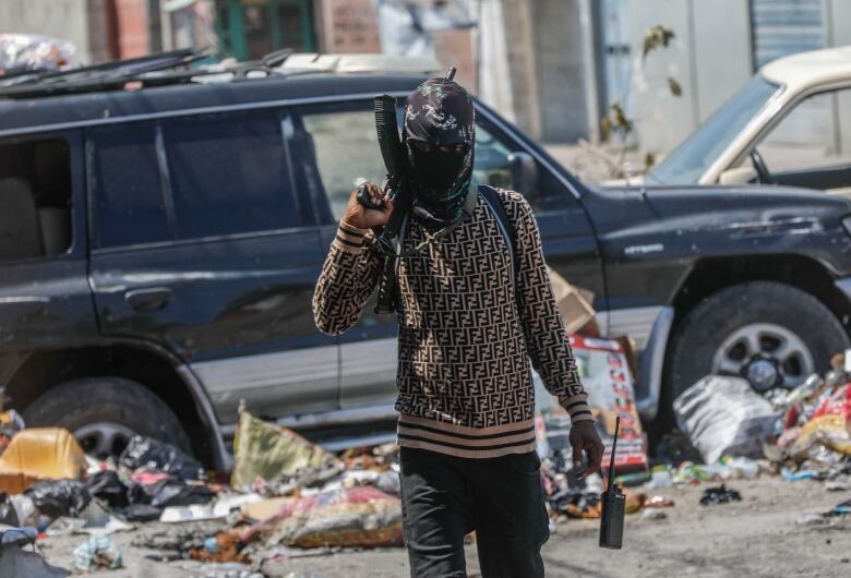 An armed gang member wearing a face mask walks down a street with a gun over his shoulder.