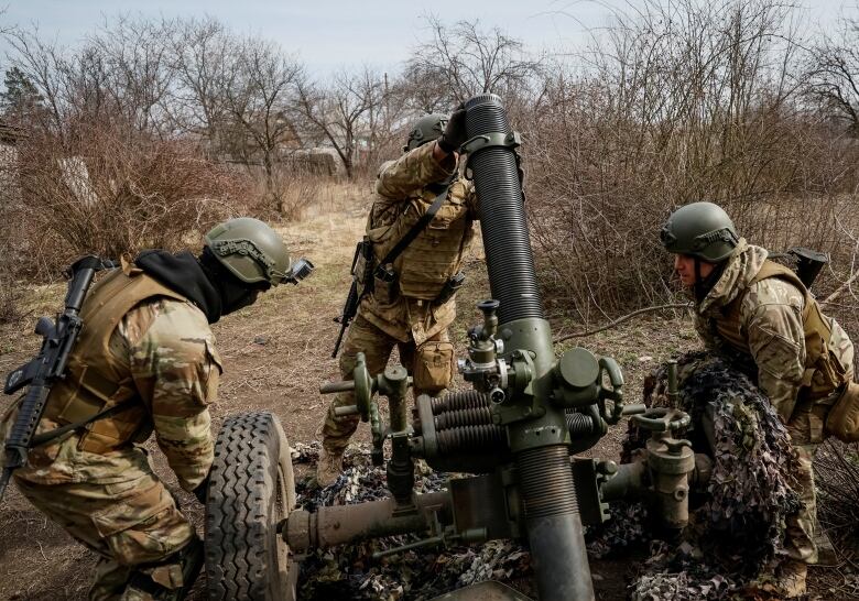 Three soldiers in camouflage near a mortar on wheels in a brown landscape.