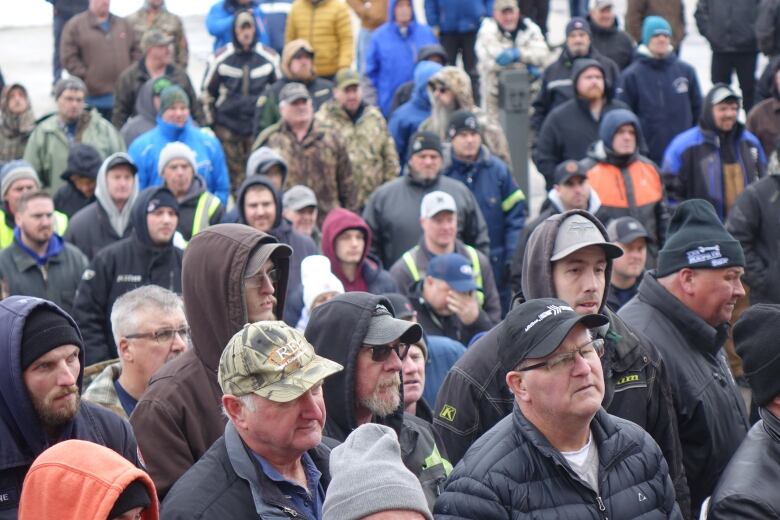 A crowd of men stand together in a parking lot listening to a speaker.