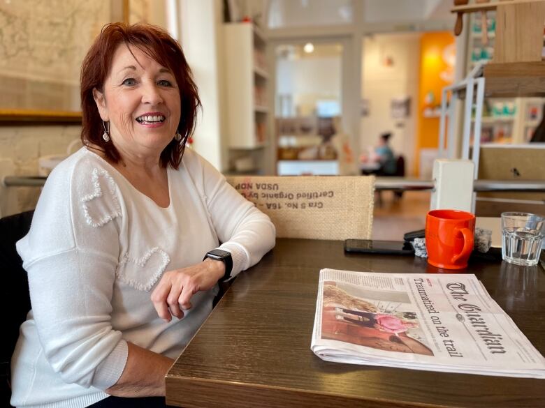 A woman in a white shirt sits at a cafe table with a newspaper in front of her.