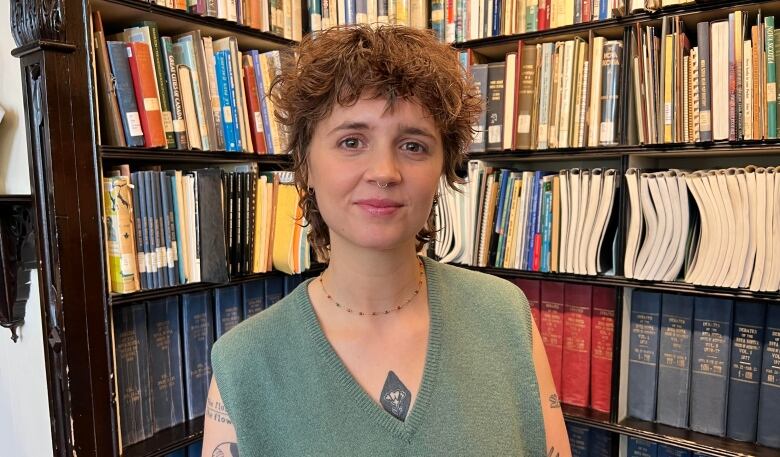 A woman with curly hair wearing a green vest, standing in front of bookshelves.