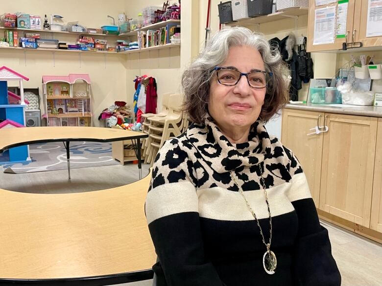 A woman with grey curly hair and glasses sits in a child's playroom.