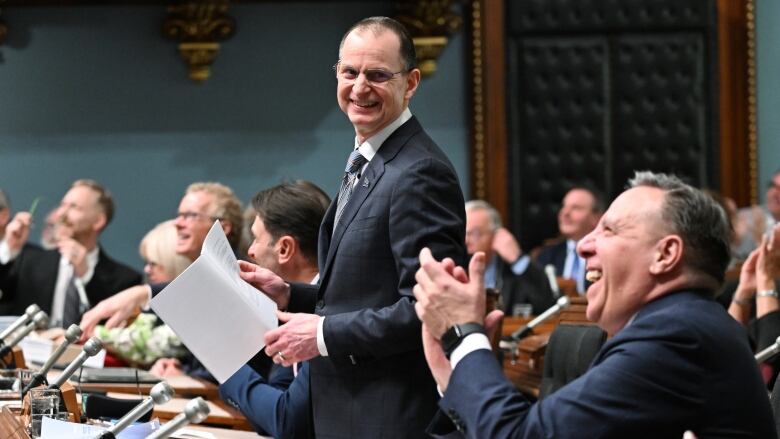 Man standing in a legislature, another man applauds in foreground.