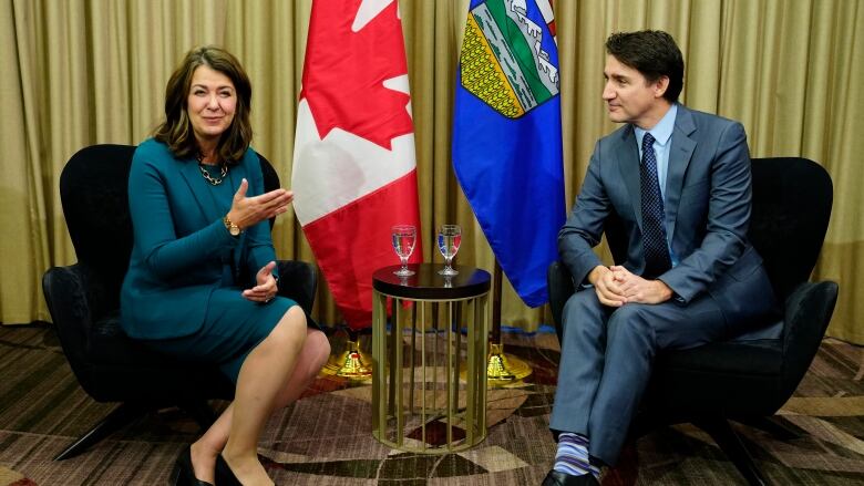 Prime Minister Justin Trudeau seated opposite Alberta Premier Danielle Smith. A small side table with water glasses sits between them with flags of Alberta and Canada on poles in the background.