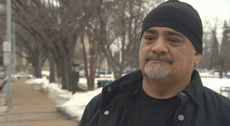 A man with a goatee is pictured on a partially snow-covered walkway wearing a black toque and a black t-shirt underneath a black jacket.