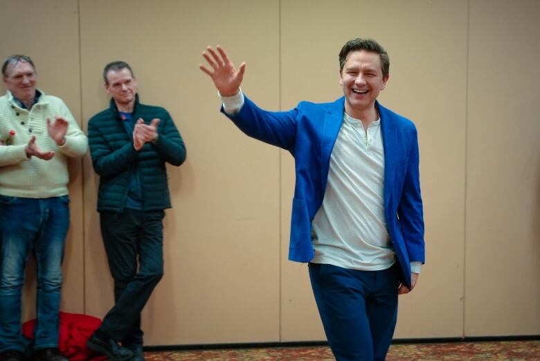 A middle-aged man with dark hair and  blue blazer smiles and waves to people off camera