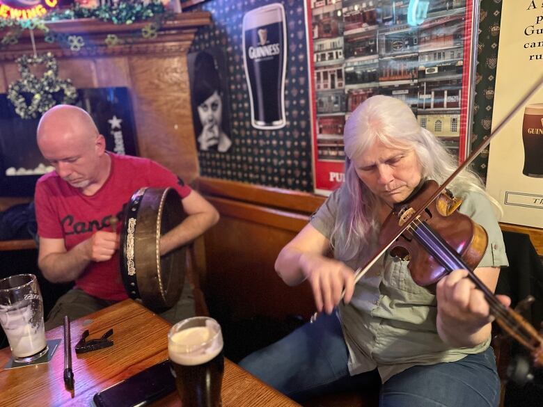 a woman plays the fiddle while a man in a red shirt plays an irish drum inside a pub