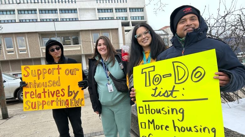 Four people pose for a photo, two of them holding signs with messages about supporting unhoused people.