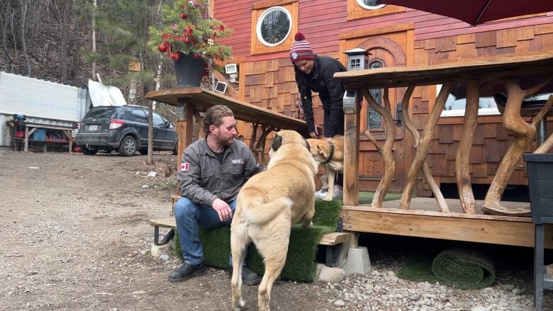 A middle aged man is sitting on the steps of the deck of his tiny home petting his large dog with his wife standing nearby petting a second dog on their wooden deck.