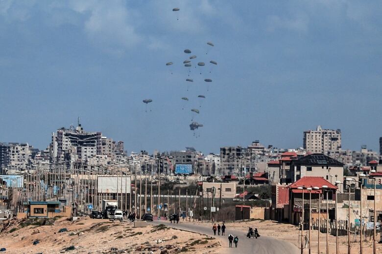 Parachutes attached to parcels of aid drop from the sky over a city with destroyed buildings.